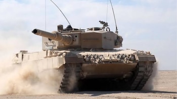 Tanque militar de color arena con cañón, torreta, escotillas y orugas; se desplaza sobre un terreno árido, levantando polvo, bajo un cielo despejado