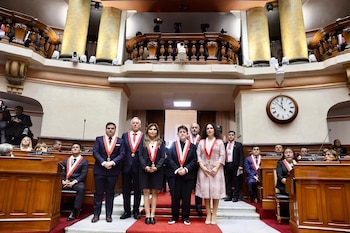 Seis personas con bandas ceremoniales sobre una alfombra roja, dos personas sentadas, una sala con columnas, balcón superior y un reloj de pared