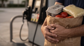 Manos de una persona que sostienen una bolsa de papel marrón con un cartón de huevos, tomates y arroz frente a un surtidor de gasolina.