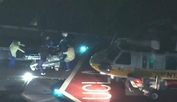 Varias personas con uniforme médico desplazan a un paciente en una camilla junto a un helicóptero sobre un helipuerto nocturno