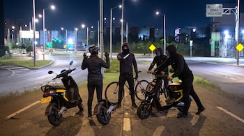 Una persona con casco y las manos en alto enfrenta a tres hombres encapuchados con un cuchillo junto a dos motocicletas en una calle.