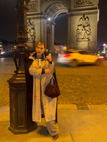 Una mujer con gabardina posa junto a una farola frente al Arco de Triunfo iluminado por la noche con un vehículo en movimiento