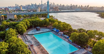 Vista aérea de una gran piscina rectangular con agua azul rodeada de árboles verdes. Un puente grande cruza un río y edificios altos se ven al fondo