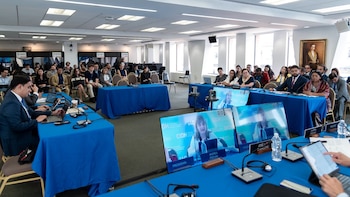 Sala de conferencia con mesas cubiertas de tela azul, pantallas mostrando videoconferencia, varias personas sentadas, micrófonos y laptops