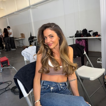 Mujer sentada con cabello largo castaño, top marrón y pantalones de mezclilla. Estudio con paredes blancas, dos personas y equipos al fondo