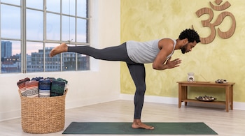 Hombre en postura de equilibrio sobre una esterilla junto a un cesto con mantas y un símbolo de Om en la pared.