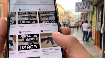 Una mano sostiene un teléfono móvil mostrando publicaciones de venta de armas de fuego en Oaxaca con imágenes de rifles. Al fondo, una calle urbana con un letrero de Cyber Café.