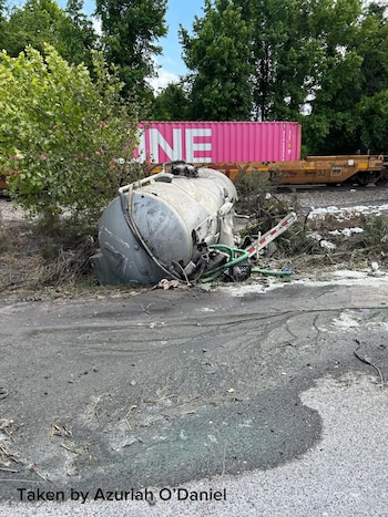 Un tanque de camión séptico volcado y gravemente dañado yace en el suelo junto a unas vías de tren, con un vagón de carga rosa en el fondo