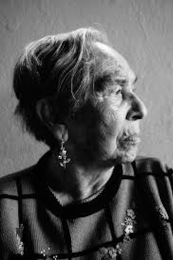 Primer plano en blanco y negro del perfil de una mujer mayor con cabello claro y arrugas faciales, usando un pendiente y un suéter estampado