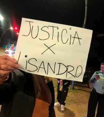 Una persona sostiene un cartel blanco con la frase "Justicia x Lisandro" escrita en letras negras. Es de noche. Hay otras personas y vehículos en el fondo