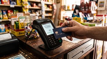 Una mano acerca una tarjeta azul a un terminal de cobro que indica un monto en pesos argentinos sobre un mostrador de madera.