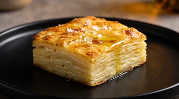 Porción cuadrada de milhojas de papa en un plato negro, con superficie dorada, escamas de sal blanca y gotas de aceite. El fondo está desenfocado.