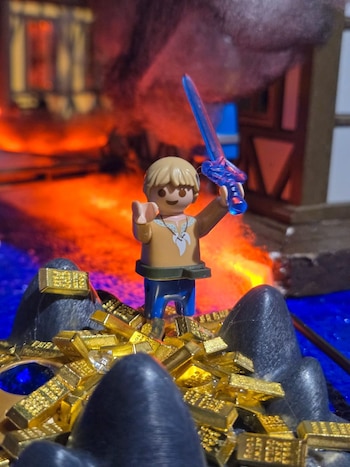 Figura de PLAYMOBIL con espada azul, de pie sobre una pila de barras de oro. Se ven formaciones rocosas oscuras y un fondo difuso naranja y azul
