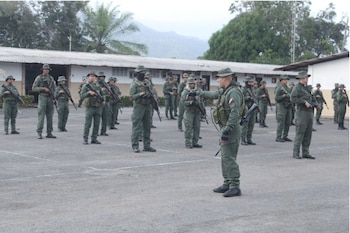 Soldados con uniformes verdes, sombreros y fusiles forman filas en un patio exterior frente a edificaciones de una sola planta