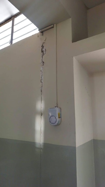 Una grieta vertical se extiende por una pared de color claro dentro de un edificio. Una ventana en la parte superior y un dispositivo blanco están visibles