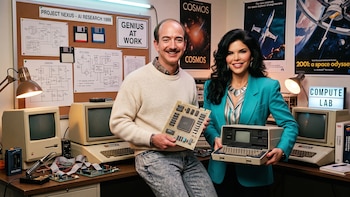 Jeff Bezos sostiene una tarjeta de circuito y Lauren Sánchez lleva una computadora portátil retro en una oficina con ordenadores antiguos