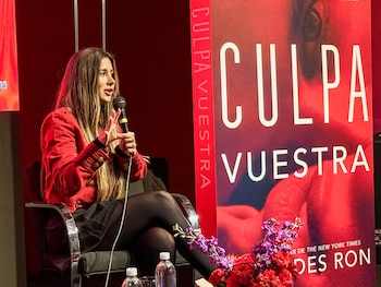 Mujer sentada, con chaqueta roja y cabello largo, hablando por un micrófono y gesticulando. Detrás, un cartel rojo con el título 'Culpa Vuestra' y flores