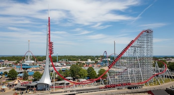 Vista aérea de una montaña rusa roja y plateada con una torre alta en un parque de atracciones. Otras atracciones, árboles, edificios y un lago son visibles bajo cielo azul.
