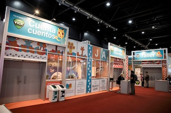 Vista de un pabellón con stands coloridos de "Cuenta Cuentos" y "Taller" en una feria. Hay algunas personas, alfombra roja y botes de reciclaje visibles