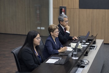 Tres personas sentadas en una mesa de conferencias con laptops y vasos de agua. Dos mujeres y un hombre con trajes oscuros. Fondo de pared de madera.