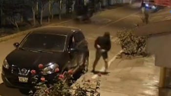 Un automóvil negro estacionado en una calle de noche junto a una persona con ropa oscura que camina por la acera