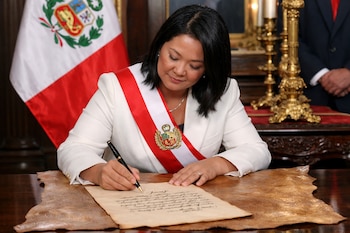 Keiko Fujimori firma un documento con una pluma negra. Viste chaqueta blanca y una banda presidencial de Perú. Detrás, la bandera de Perú y un candelabro dorado. El documento está sobre un cuero de becerro.