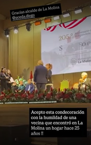 Un escenario con personas, un hombre de espalda que entrega un reconocimiento a una mujer, una pantalla gigante, luces, un podio floral