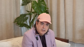 Hombre con gorra rosa y chaqueta lila sentado, un micrófono prendido en la solapa, cortina clara y planta verde de hojas grandes al fondo