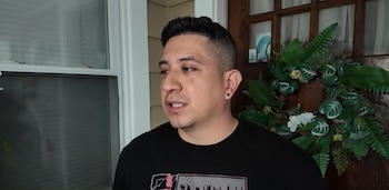 Un hombre de cabello oscuro y camiseta negra aparece de medio cuerpo frente a una ventana con persianas y una puerta decorada con una corona de hojas