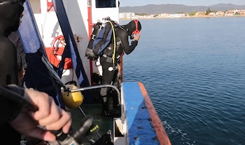 Dos personas con trajes de neopreno en una embarcación. Un buceador con equipo completo se inclina hacia el agua. Detrás, costa con edificios y montañas