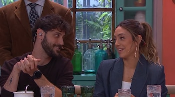 Un hombre con barba y una mujer con el cabello recogido conversan en una mesa frente a varios vasos de vidrio.