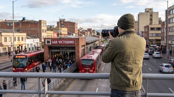 Un hombre de espaldas toma una fotografía con una cámara hacia la estación San Victorino de TransMilenio y la avenida con autobuses rojos.