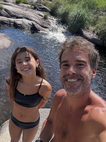 Hombre y niña sonriendo a cámara en primer plano. El hombre sin camiseta y la niña con bikini negro. Detrás, un río con rocas y una pequeña cascada