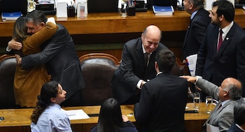 En un recinto legislativo, un hombre calvo con traje oscuro sonríe y saluda de mano. Cerca, un hombre abraza a una mujer. Otros individuos están presentes