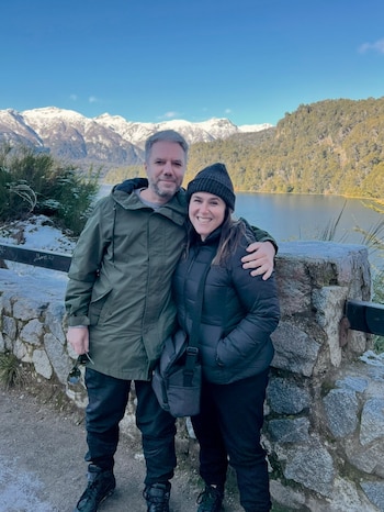 Hombre y mujer sonríen, vestidos con ropa de abrigo, posando junto a una baranda de piedra con nieve, lago, montañas nevadas y cielo azul de fondo