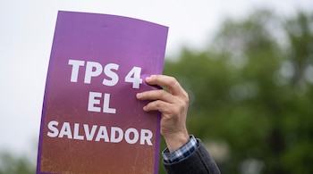 Mano de persona sosteniendo un cartel púrpura con las palabras 'TPS 4 EL SALVADOR' en letras blancas. Fondo desenfocado de cielo y árboles.
