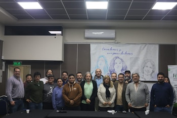 Veinte adultos, en su mayoría hombres, posan sonrientes en una sala. Detrás, un cartel blanco con dibujos de mujeres y el texto "Creadoras y emprendedoras"