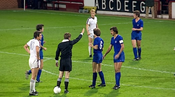 Un árbitro de negro levanta el brazo junto al balón frente a varios futbolistas con uniformes azules y blancos en la cancha.