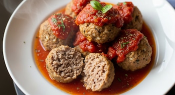 Un plato blanco contiene varias albóndigas de arroz y carne en salsa de tomate, decoradas con hierbas. Una de ellas está cortada revelando su interior.