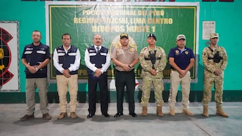 Siete hombres con uniformes policiales, militares y chalecos institucionales posan de pie delante de una pared verde con un cartel oficial