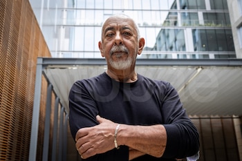 Primer plano de Leonardo Padura, hombre con barba y cabello canoso, camiseta negra, brazos cruzados, con un edificio de arquitectura moderna al fondo