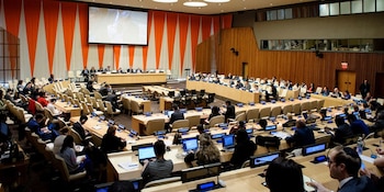 Vista aérea de un amplio salón de conferencias con mesas y sillas ocupadas por numerosos delegados, frente a un estrado y una pantalla grande proyectando una imagen