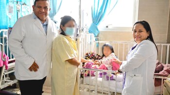 Un médico y una médica de bata blanca, una mujer con mascarilla y una niña en una cuna. Se ven cortinas azules de hospital y equipos médicos