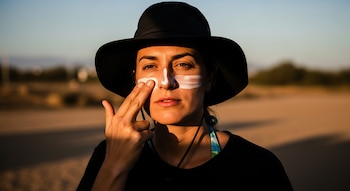 Primer plano de una mujer con sombrero oscuro aplicando protector solar blanco en su nariz y pómulos con su dedo índice, bajo la intensa luz del sol en un exterior.