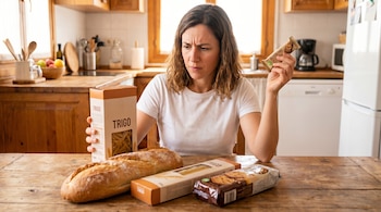 Una mujer sentada a una mesa de madera en una cocina, sosteniendo una barrita con la etiqueta "gluten free" y mirando un paquete de pasta de trigo, con pan y galletas cerca.