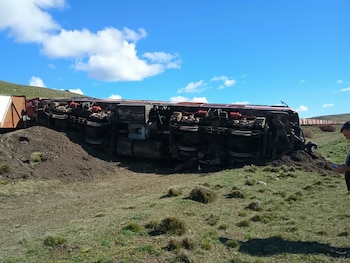 Un vagón de tren volcado de costado sobre un terreno de hierba y tierra, bajo un cielo despejado con nubes