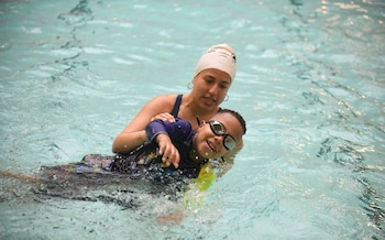 Una mujer con gorro de natación beige y un niño con gafas nadando en una piscina. La mujer sostiene al niño, quien sonríe y chapotea agua