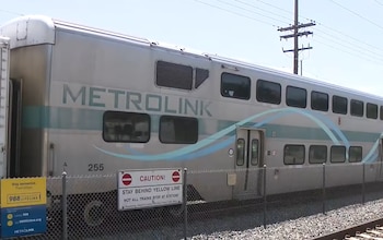 Tren Metrolink plateado con logo verde, número 255, ventanas. Hay una valla metálica, carteles de advertencia y vías del tren bajo cielo claro