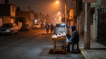 Un joven sentado en un banquillo junto a una mesa con dulces en una calle nocturna. Se observan coches estacionados, personas caminando y un letrero que dice Edomex-Toluca.