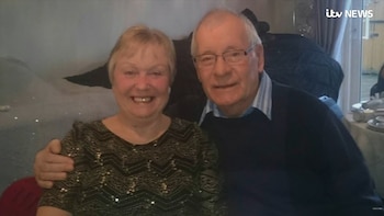 Un hombre y una mujer de edad avanzada posan sonrientes, con el hombre usando gafas y la mujer un top estampado, en un ambiente interior